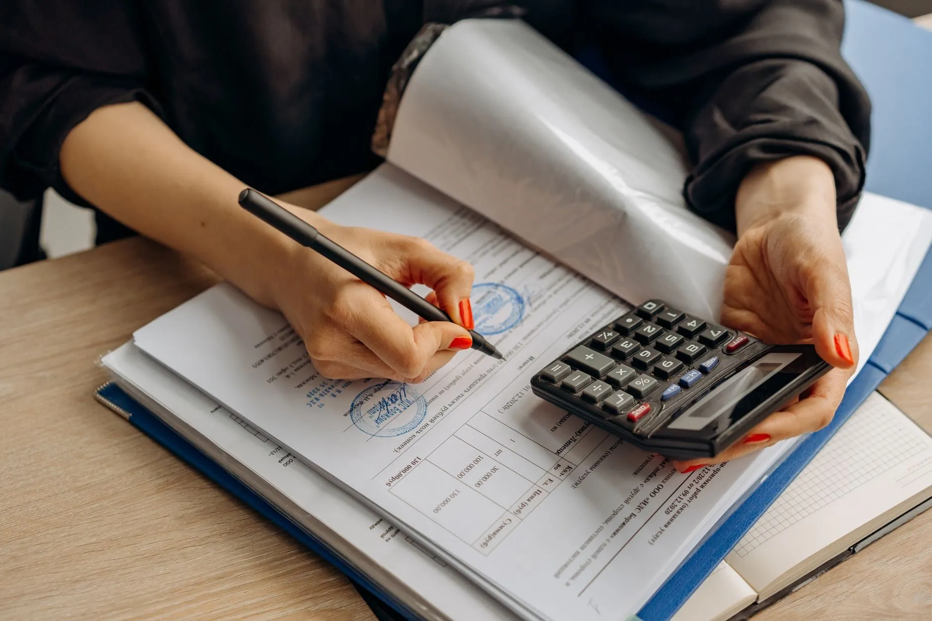An accountant signing a stamped document while holding a calculator over a blue folder